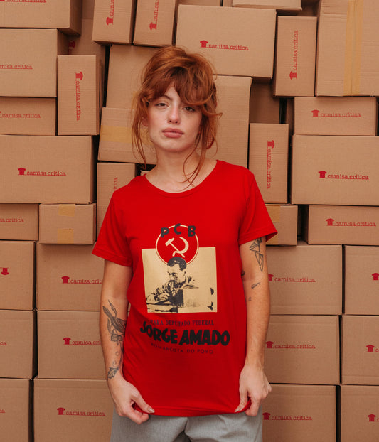 Mulher vestindo Camiseta Baby Look Estampa: Acima a insígnia do PCB com a foice e o martelo. No meio uma foto de Jorge Amado fumando e escrevendo numa maquina de escrever. Abaixo está escrito: Para deputado federal Jorge Amado Romancista do Povo