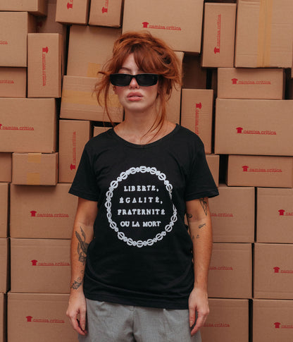 Mulher vestindo Camiseta Baby Look Estampa:  insígnia da Revolução Francesa. texto com os dizeres em francês: Liberte, Egalite, Fraternite ou La Mort. Truz: Liberdade, Igualdade, Fraternidade ou a Morte. Fios transados circulam o texto.